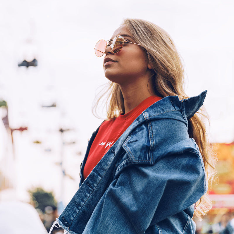 Junge Frau mit langen blonden Haaren trägt rote Shirt, Jeansjacke und runde Sonnenbrille, steht selbstbewusst vor urbaner Kulisse mit Riesenrad im Hintergrund – moderner Streetstyle-Look, ideal inszeniert für minimalistische Accessoires und Lifestyle-Fotografie.