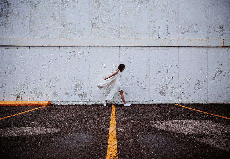 Frau in weißem, fließenden Kleid läuft dynamisch über einen leeren Parkplatz mit gelber Markierung vor verwitterter Betonwand – minimalistischer, urbaner Look für moderne Mode- und Lifestyle-Inszenierungen.