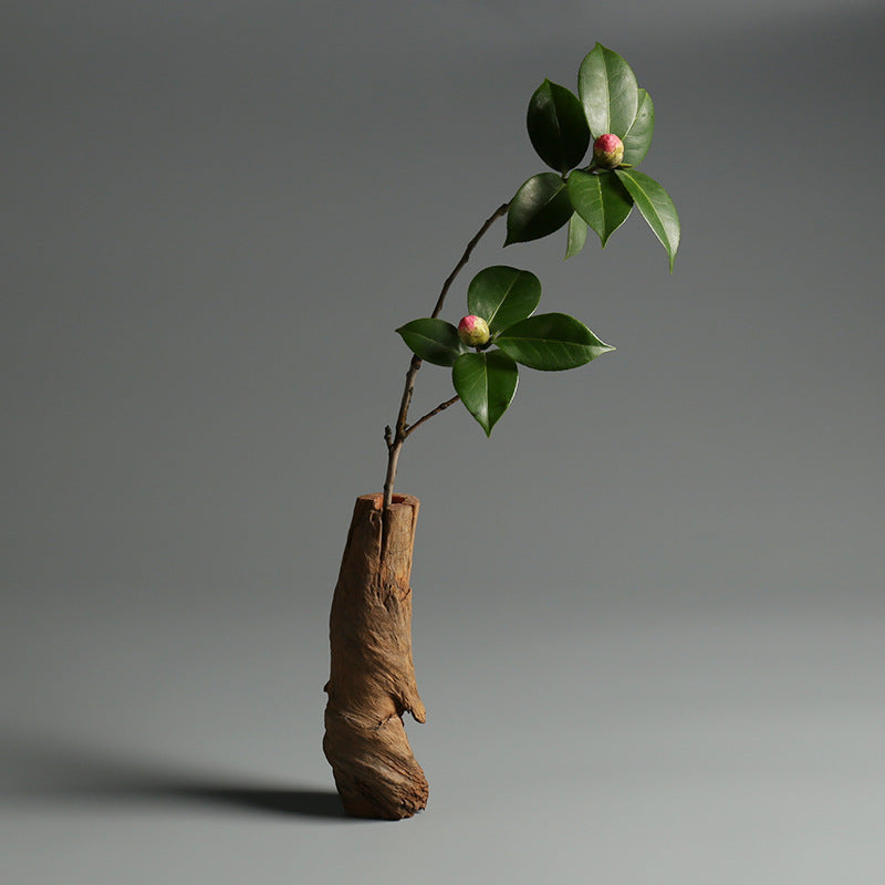 Ein minimalistisches Arrangement mit einem Zweig mit grünen Blättern und rosa Knospen in einer rustikalen, zylindrischen Holzvase vor einem grauen Hintergrund.