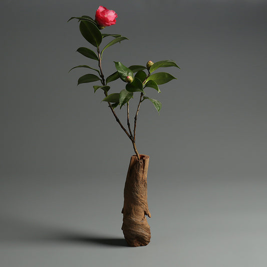 Eine rosa Blume mit grünen Blättern in einer minimalistischen Holzvase vor einem schlichten grauen Hintergrund.