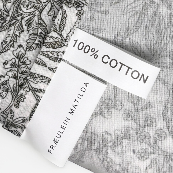 Detailaufnahme eines Stoffetiketts mit der Aufschrift „100 % Cotton“ und Markenlabel „FRÄULEIN MATILDA“ auf botanisch bedrucktem Naturstoff – hochwertige Baumwolle für stilvolle Wohnaccessoires mit natürlichem Charakter.